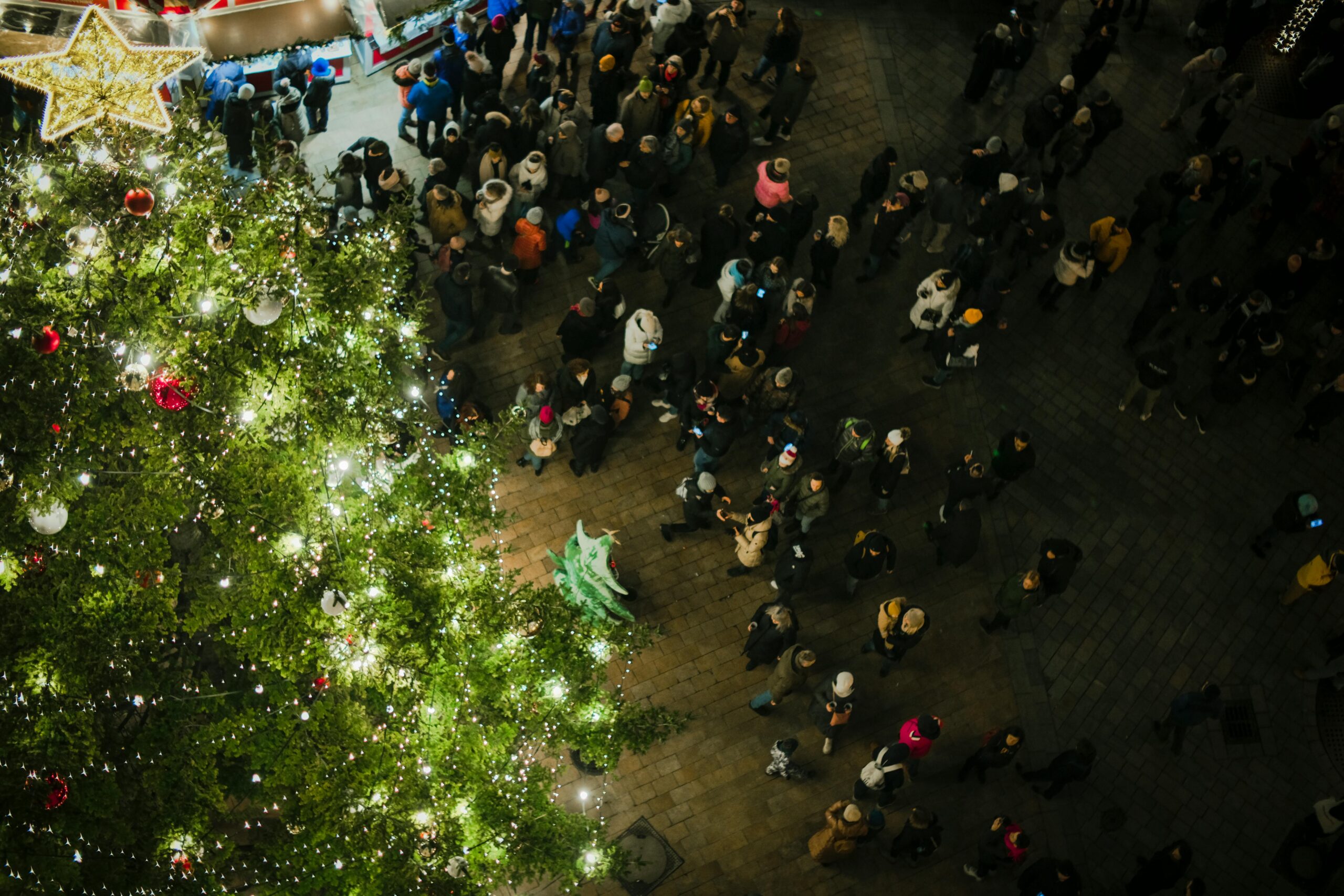 Vista dall’alto di una piazza illuminata per Natale con albero, bancarella e persone che passeggiano di sera